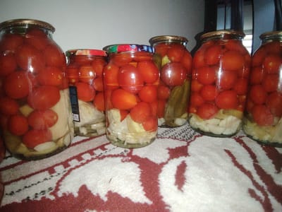 Six glass jars filled with preserved tomatoes and other vegetables, possibly pickles or cabbage, are arranged on a patterned tablecloth. The jars are of varying sizes, and several have decorative lids. The background is dimly lit, with a hint of wooden furniture or shelving. The scene appears to be in a domestic setting, likely a kitchen or pantry, in the city of Pitnak, Uzbekistan. There are no people visible in the image.