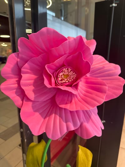 A vibrant pink, large-scale artificial flower dominates the foreground of the image. The flower has many layers of ruffled petals, with a detailed center featuring yellow and pink stamen-like structures. The petals are a bright, saturated pink and appear to be made of a soft, matte material. Sunlight casts shadows across the petals, highlighting their texture and form.

In the background, out of focus, are architectural elements that suggest an indoor, possibly commercial or public space. There are dark vertical structures, reflective surfaces, and lights that appear to be ceiling fixtures. Some beige tiled flooring is also visible. There is no indication of the city of Slitu, Norway, within the image itself. No people are visible in the frame. Two large, yellow artificial leaves extend from the bottom of the frame, connected to a green stem.