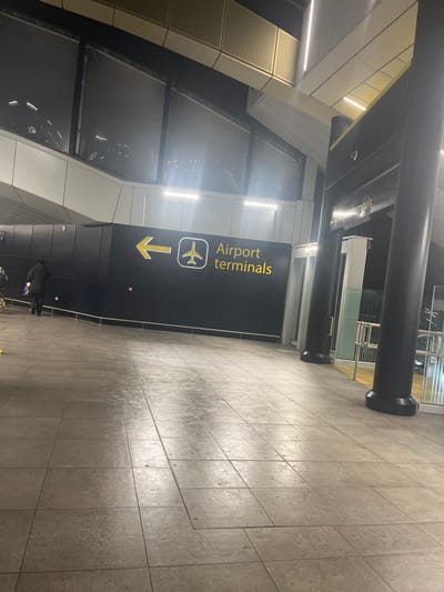 The image displays an indoor scene, likely within an airport terminal or an adjacent connecting area, characterized by modern architecture and artificial lighting. The perspective is rotated 90 degrees clockwise from a standard horizontal view, with the floor running vertically on the right side of the frame.
A prominent black wall panel features clear white and yellow signage. The text "Airport terminals" is visible, accompanied by a white airplane icon above a yellow arrow pointing to the right (if the image were correctly oriented horizontally). This sign serves as a directional guide for travelers.
The setting includes a tiled floor made of light grey, rectangular slabs, showing reflections from the overhead lighting. Large, dark cylindrical pillars support the ceiling structure, which incorporates linear light fixtures emitting bright, white light. The walls are a mix of dark panels and lighter, possibly metallic or textured, panels. Another overhead beam, running parallel to the "Airport terminals" sign, has some reflective, indistinct text on its underside that is illegible due to glare and blur, but it might indicate "South Terminal" or similar.
A few indistinct human figures are visible in the distance, walking away from the viewer, near the "Airport terminals" sign. One figure appears to be carrying an item. The artificial illumination suggests either evening or night, or simply an interior space where natural light is not a dominant factor. The overall atmosphere appears functional and quiet, consistent with a transportation hub in Horley, United Kingdom.