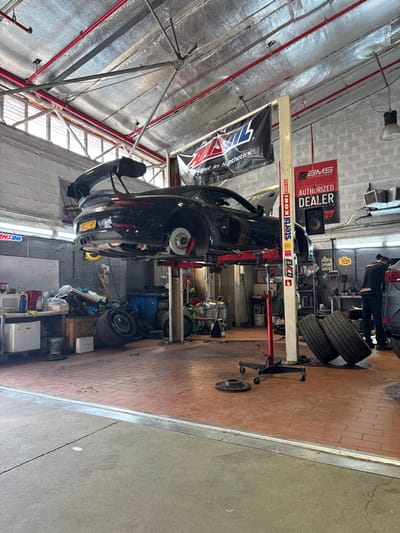 The image depicts an interior scene of an automotive repair shop in Azor, Israel. A black Porsche 911 GT3, displaying Israeli license plate 532-27-501, is centrally positioned and elevated on a red two-post hydraulic lift, bearing "Stertil Koni 5000 kg" branding. All four wheels are removed from the vehicle, exposing brake rotors and red calipers on each hub. Two loose car wheels with tires are propped against the right pillar of the lift. Two individual brake rotors lie on the reddish-brown tiled floor in the foreground.
The shop's ceiling is insulated with metallic foil material, featuring exposed red piping and overhead windows. White concrete block walls are visible, displaying a black "AMSOIL The First in Synthetics" banner and a red "AMS PERFORMANCE AUTHORIZED DEALER" banner. On the left side, a white workbench unit holds a microwave, bottles, and other items, with a cluttered storage area behind it containing additional loose wheels and equipment. In the far right background, two individuals, dressed in dark attire, are partially visible near a silver-grey vehicle; one is bent over its front section, while the other stands upright nearby. The immediate foreground shows a grey concrete floor transitioning into the tiled shop area.