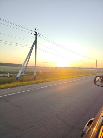The image captures a roadside view at sunset in Rastunovo, Russia. A paved road with a white line marking the edge stretches into the distance, blurred by motion. To the left of the road, a vast field of tall grass and crops is illuminated by the setting sun, which is low on the horizon and casts a golden glow. A large, angled power line pole with multiple wires runs parallel to the road, with another smaller pole visible further down the field. A white fence runs along the edge of the field, and a line of trees forms the horizon. In the lower right corner of the image, a car's side mirror is visible, reflecting the golden light and a hint of the car's interior. The overall scene depicts a rural landscape under a clear, pale sky transitioning into dusk.