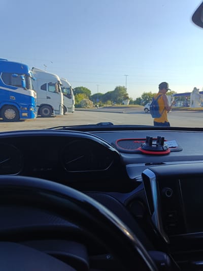 The image is taken from inside a vehicle, looking out through the windshield at a parking lot with several large trucks. In the foreground, the dashboard of the vehicle is visible, including part of the steering wheel and a phone holder. The dashboard contains various gauges and buttons, including what appears to be a speedometer.
Outside the vehicle, three trucks are parked: a blue and white truck on the left, and two white trucks to its right. Beyond the trucks, there is a paved area, some greenery, and a clear blue sky. A person wearing a yellow tank top and a blue backpack is walking away from the camera, holding a phone. There are also some buildings and structures in the background, including what looks like a gas station or service area with a large white statue.
The lighting suggests it is daytime, likely morning or late afternoon, given the angle of the sun and the long shadows. The sky is clear, indicating fair weather. The scene appears to be a truck stop or a rest area, possibly near a highway or a service road.
Visible text includes the words "SRC" and "MENU" on the vehicle's dashboard. There is also some faint text on a slip of paper attached to the phone holder, which is too blurry to read clearly but appears to be a date. The location context provided is Riccione, Italy. The overall impression is a typical roadside scene during a journey.
