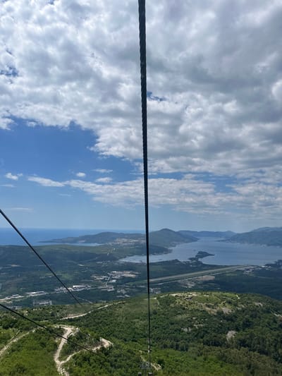 The image captures a view from a cable car traversing above Mirac, Montenegro. The scene is oriented such that the sky, partially covered with white clouds against a blue backdrop, occupies the left side. The landscape on the right encompasses a mountain range densely covered with green vegetation, a winding dirt road visible on the mountainside, and the coastline of the Bay of Kotor. In the middle ground, the bay extends towards the horizon, dotted with small islands and framed by land masses. A highway traces part of the shoreline. Foreground cable lines from the cable car are visible.