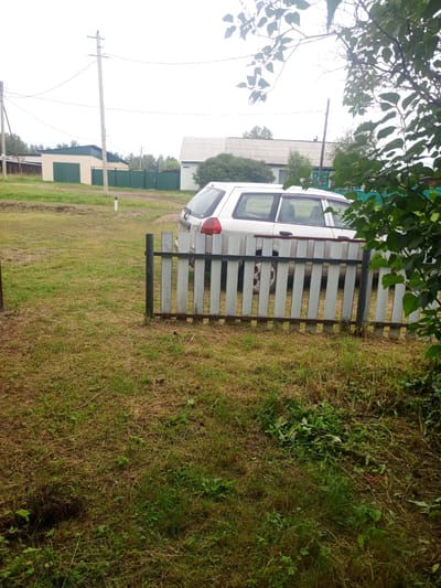 A white station wagon is parked in front of a light-colored wooden fence. The car is positioned on the right side of the frame, with its rear partially visible. The fence runs horizontally across the middle of the image. Beyond the fence, a grassy area stretches towards several buildings in the background. One building is a light beige structure with a green roof and green garage doors. Another building, further to the right, appears to be a white house with a grey roof. Electrical poles and wires are visible in the sky above the buildings. Trees and foliage are scattered throughout the scene, with some branches and leaves obscuring parts of the sky and buildings on the right side. The ground is covered in dry grass and some green patches of vegetation. The scene depicts a rural or suburban setting in Kuytun, Russia, with a typical residential landscape.