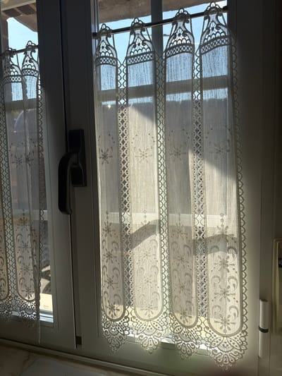 The image shows a close-up view of a window with white lace curtains hanging. The window appears to be part of a building, and sunlight streams through the curtains, creating patterns of light and shadow. The curtains have a decorative, intricate design with floral motifs. A dark-colored window handle is visible on the left side of the frame. The overall impression is one of domestic interior, with the window providing a view to the outside, though the external environment is not clearly discernible. The location is identified as Arcahueja, Spain.