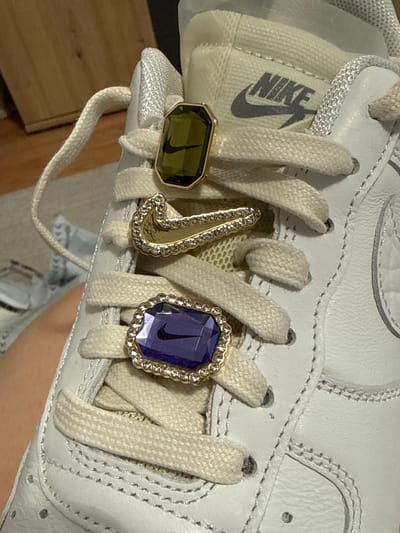 A close-up shot captures the top section of a white Nike sneaker, adorned with several decorative elements. The shoe itself appears to be made of white leather or synthetic material, featuring subtle stitching details along the laces. Off-white, flat woven shoelaces are threaded through the eyelets.
Three distinct, jewel-like lace charms (dubraes) are prominently displayed on the laces. The leftmost charm is a rectangular, olive green facet-cut gem set in a gold-colored frame, with a small black Nike swoosh logo embedded within it. The middle charm is a stylized gold-colored "J" shape, encrusted with numerous small clear rhinestones or crystals. The rightmost charm is an octagonal or cushion-cut deep purple facet-cut gem, also framed in gold-colored metal and surrounded by small clear rhinestones, featuring another black Nike swoosh logo inside.
Visible on the shoe's tongue, behind the laces, is the iconic "NIKE" text logo in black. The background of the image is softly blurred, showing hints of a warm-toned, possibly wooden, surface. The overall impression is a detailed showcase of personalized footwear accessories. The image does not contain any visual cues pertaining to the specified location of Hainburg an der Donau, Austria.