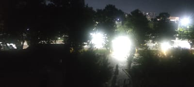 The image depicts a nighttime street scene in Andijan City, Uzbekistan, characterized by strong artificial lighting and dark shadows cast by trees. The foreground is dominated by the dark silhouettes of trees and foliage. In the midground, a pathway or road is visible, illuminated by intensely bright, diffused lights that appear to be streetlights or vehicle headlights, creating a blown-out white effect in the center of the image. These lights are partially obscured by trees, suggesting a park or tree-lined street. In the background, faint outlines of buildings with illuminated windows can be seen, indicative of urban development. There are two indistinct human figures walking on the illuminated path in the middle ground, their forms elongated by the low-angle lighting. The overall atmosphere is nocturnal and somewhat hazy due to the intense light sources.