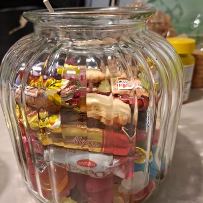 The image shows a glass jar filled with various candies. The jar has a fluted design, and the light creates reflections and refractions through the glass and candies. The candies are individually wrapped in colorful foil, with some wrappers showing text. Visible text includes "Mifare", "Bucuria", and potentially other brand names in Cyrillic script. There are also some unwrapped jelly-like candies in shades of red, pink, and blue. In the background, out of focus, are some yellow objects, possibly lids or containers, and a green textured surface. The lighting suggests an indoor setting, likely a kitchen or pantry, with the light source coming from the top. The overall scene conveys a sense of abundance and variety of sweets. There are no people or animals visible in the image. The context of Bösel, Germany is provided, but there are no specific visual cues in the image that directly point to this location.
