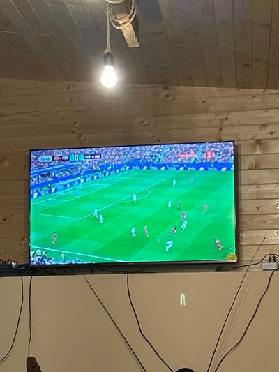The image shows a television screen displaying a soccer match. The screen is mounted on a wooden wall. On the screen, players in white and red uniforms are visible on a green field. The score board indicates the time as 10:48. There are several logos of "VISA" visible on the screen. In the foreground, electrical cables are seen hanging down from the ceiling. The scene appears to be indoors, possibly in a living room or a recreational area, with a rustic wooden interior.