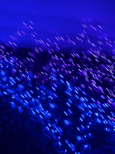 An abstract, dark blue image features streaks of light resembling rain or motion blur. These illuminated lines are mostly blue, with some hints of purple and pink. The left side is predominantly a solid blue, creating a contrast with the clustered streaks on the right. The scene is dark and blurry, the texture gives the impression of a night scene with fast-moving lights.