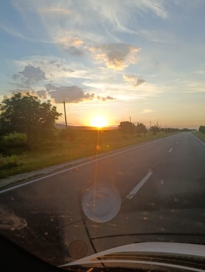 The image captures a picturesque sunset over a rural landscape, viewed from the perspective of someone inside a vehicle. The foreground shows the car's dashboard and windshield, with reflections and some dust particles visible. The main subject is a paved road stretching into the distance, bordered by grassy fields and scattered trees. A bright, radiant sun is setting on the left side of the horizon, casting a warm, golden glow over the scene and creating lens flares. The sky is a canvas of soft blues and oranges, with scattered clouds tinged by the setting sun. A lone utility pole stands by the roadside, suggesting the presence of power lines. The overall atmosphere is serene and tranquil, evoking a sense of journey or travel at the end of the day. The time of day is clearly sunset. The weather appears clear with some clouds. No text is visible in the image.