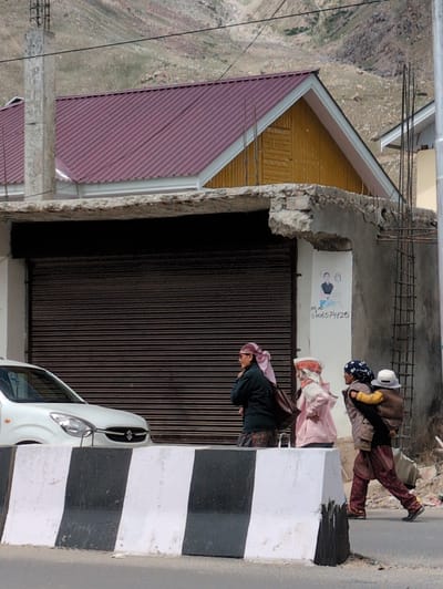 In Padum, India, a group of individuals walks along a street. In the foreground, a white car is partially visible behind a black and white striped concrete barrier. Behind the barrier, three people are walking from left to right. The first person on the left is wearing a dark jacket and a pink headscarf. The second person is wearing a light pink hooded jacket and a white and red patterned headscarf. The third person is carrying a child on their back. The child is wearing a white hat. This person is wearing a dark vest over a patterned outfit and a dark blue headscarf. The background shows a building with a maroon roof and yellow siding, and further back, a mountainous landscape.