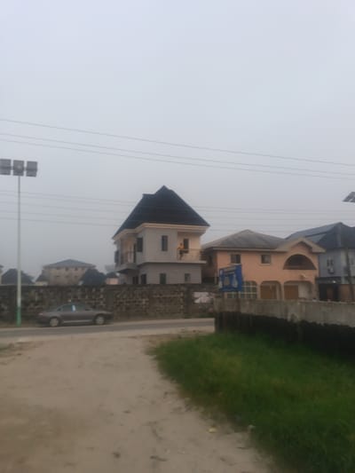 A hazy day scene features a sandy road leading to a row of houses. A gray car is parked near a dark brick wall in front of a two-story house with a dark roof. Adjacent to it is a peach-colored house with an arched window detail and a sign in front. Grass grows along the edge of the road, and utility lines stretch across the sky. A tall pole with multiple lights stands on the left.