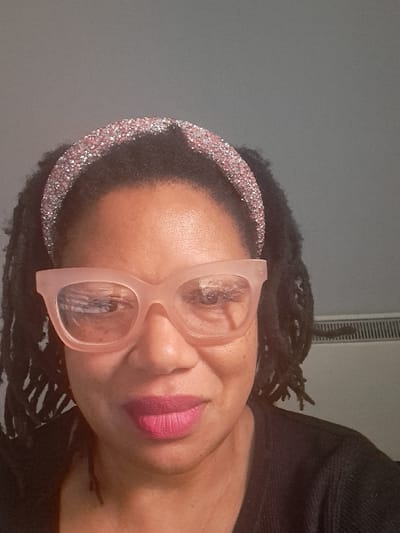 The image displays an indoor headshot of an adult female with dark skin, directly facing the camera. Her dark hair is styled in locs, and she wears a thick, sparkling pink or rose gold headband. She is wearing large, light pink-framed, cat-eye-style glasses, with reflections visible on the lenses. Her lips are colored with bright pink lipstick. She wears a dark, textured garment, likely black. The background features a plain, light grey wall. A white panel radiator with vertical fins is partially visible on the right side of the frame, behind the subject's right shoulder. The precise location within Stevenage, United Kingdom, cannot be determined from the visual information provided.