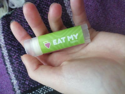 A close-up shot shows a hand holding a tube of lip balm. The lip balm tube is green and has the text "EAT MY balm" written in white. There is also a small pink strawberry graphic on the tube. The hand is resting on a purple, textured fabric, possibly a blanket or scarf. The background is slightly blurred, but appears to be indoors with soft lighting. There is no specific indication of the location within Шарпілаўскі сельскі Савет, Belarus.