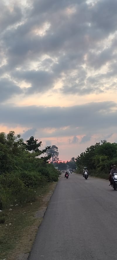 A vertical, eye-level shot captures a scenic road in Blangmajron, Indonesia, at sunset. The sky is filled with dramatic, cloudy formations in shades of gray, with hints of pink and orange peeking through, indicating the setting sun. The road stretches into the distance, flanked by lush green foliage on either side.
Several people are riding motorcycles on the road. In the foreground, on the right side of the road, a motorcycle carries two people, with the passenger partially visible. Further down the road, more motorcycles are seen, with individuals riding them. One motorcycle has a rider with a green helmet. The scene appears to be a typical evening commute or a leisurely ride through the countryside.
The overall atmosphere is serene and peaceful, amplified by the soft glow of the sunset. The abundance of greenery suggests a rural or semi-rural setting. There is no visible text in the image. The weather is overcast but not raining, with the clouds creating a visually interesting backdrop for the sunset. The time of day is clearly evening, as evidenced by the sunset colors in the sky.