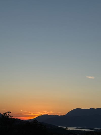 The image shows a sunset over the Bay of Kotor, with the mountains of Pelinovo, Montenegro in the foreground. The sky is a gradient of blue and orange, with a few wispy clouds. The mountains are silhouetted against the colorful sky, and a body of water is visible in the distance. The overall scene is peaceful and serene.