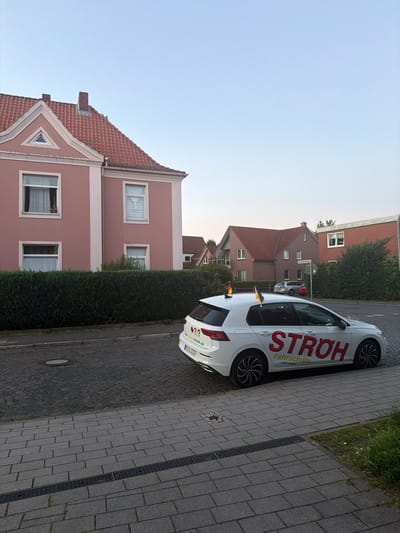 A white Volkswagen Golf, a driving school vehicle for STROH Fahrschule, is parked on a cobblestone street in the city of Nienburg/Weser, Germany. The car has German flags on its front pillars and the driving school's branding, "STROH Fahrschule ... und mehr," in large red letters on its side. The license plate is visible and reads "NI-LW 3333".

The car is positioned in front of a pink-colored residential building with a red-tiled roof, partially obscured by a dense green hedge. To the right of this building, another house with a red-tiled roof is visible, and further to the right is a brick building with a flat roof and white-framed windows. The street is paved with cobblestones, and a sidewalk made of grey paving stones with a drainage grate runs in front of the parked car. The sky is clear and blue, indicating daytime. The overall impression is a quiet residential street scene.