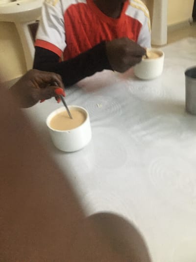 Two white cups containing beige-colored liquids sit on a white surface. A person wearing a red and white shirt and black sleeve holds a silver spoon in their hand, stirring one of the cups. The person's hand has orange nail polish. The image is taken from an overhead perspective and is slightly blurred, with some of the person's body visible in the lower right corner.
