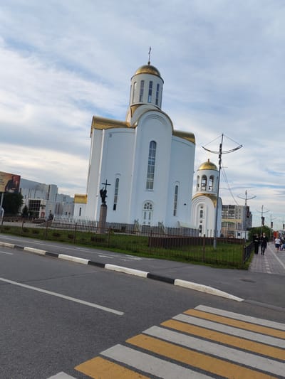 The image shows the Transfiguration Cathedral in Kurgan, Russia, from a street-level perspective. The white cathedral features gold domes and a cross on its main dome. To the left of the cathedral, a statue of an angel holding a cross stands on a pedestal. A black metal fence separates the cathedral grounds from the sidewalk and road.

In the foreground, a pedestrian crosswalk with yellow and white stripes is visible on the asphalt road. The sidewalk adjacent to the road has people walking, including a couple dressed in dark clothing and a woman in a light-colored top and shorts. Behind the cathedral, modern buildings and a billboard are visible. The sky is partly cloudy with a mix of blue sky and white clouds. The scene appears to be in an urban environment during daylight hours.