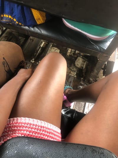 The image depicts the interior of a vehicle, viewed from a first-person perspective, looking downwards and forward. The scene is located in Ajah, Nigeria.
In the immediate foreground, the photographer's legs, with a dark skin tone, are visible. The left leg has a dark, irregularly shaped mark or stain on its inner thigh. The individual wears light-colored, horizontally striped knit shorts, predominantly red and white. A black, textured seat surface is present in the extreme foreground, partially obscured by the legs.
Further into the midground, a dark metal structural bar runs horizontally across the frame. Below this bar, the vehicle's floor is visible, appearing worn, dirty, and wet with dark, reflective patches. To the right of the photographer's legs, the lower leg and right foot of another individual, also with a dark skin tone, are visible. This foot wears a pink sandal with a light blue/teal strap. Another black, textured seat is partially visible to the right of this person's leg.
In the upper left quadrant, a blue bag with yellow circular and wave-like patterns is partially visible, resting on a surface. In the upper right quadrant, a portion of a green and white shoe is visible, potentially belonging to a third passenger or placed on an adjacent seat. The overall environment suggests an aged or utilitarian mode of transport, possibly public, given the exposed structure and condition of the floor.
