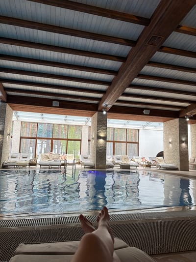 The image presents an indoor swimming pool area, likely within a luxurious spa or hotel setting in Kalinovo, Russia. The perspective is tilted, showcasing architectural features of the establishment.

On the left side, a striking ceiling or high wall is visible, constructed with light-colored, possibly white or pale wood, panels interspersed with prominent dark brown wooden beams. These beams run diagonally, creating an appealing pattern. A notable detail is a carved square design embedded in one of the horizontal wooden beams.

Towards the center of the frame, large, textured pillars or walls, made of light-colored stone-like material, stand between the main pool area and expansive windows. Recessed lighting fixtures are integrated into both the stone pillars and the wooden beams. Through the large windows, bright daylight illuminates the interior, revealing lush green foliage outdoors, suggestive of a natural landscape or garden.

The right side of the image is dominated by a clear, blue-tinted swimming pool with gentle ripples on its surface. The water reflects the intricate ceiling structure and the outside greenery. Along the far side of the pool, several modern lounge chairs, each draped with a white towel or cushion, are arranged for relaxation. Reflections in the water hint at people using these chairs, with one person visibly relaxing on a lounge chair. In the immediate foreground on the right, a person's leg and foot are visible, positioned near the pool's edge, indicating relaxation or an imminent entry into the water. A textured, patterned surface runs along the pool's side.

The scene conveys a sense of tranquility and luxury, accentuated by the architectural design, natural light, and the clear pool water. There is no visible text in the image.