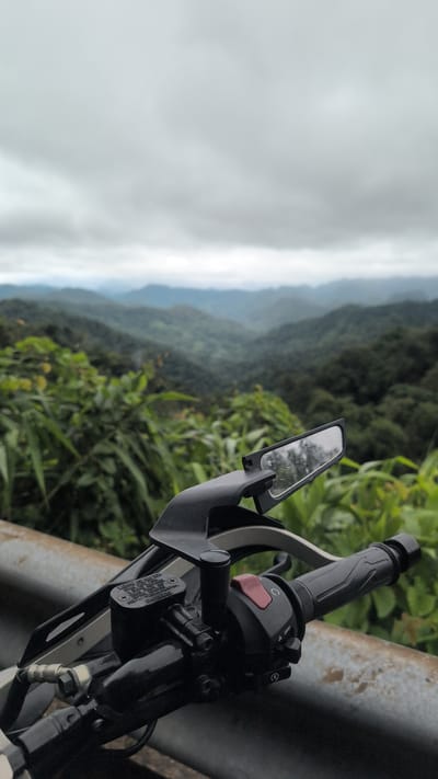 This is a photograph taken from a motorcycle, looking out over a mountainous landscape under a cloudy sky. The foreground is dominated by the motorcycle's handlebars, including a side mirror reflecting the sky and trees, a brake fluid reservoir, and control switches. The middle ground features lush green vegetation, possibly a roadside planting. The background reveals a series of rolling, tree-covered hills fading into the distance under an overcast sky. The image appears to be taken in Thượng Trạch Commune, Vietnam, a region known for its mountainous terrain and dense forests. There are no people visible in the image.