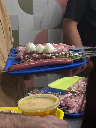 The image portrays a food preparation scene in Azor, Israel, featuring raw meats arranged on skewers for grilling. Two individuals collaboratively hold a rectangular blue plastic tray positioned centrally in the frame.
The blue tray contains multiple metal skewers. Several skewers hold light-pink raw meat chunks, likely poultry or pork. Other skewers contain a mixture of darker ground meat, finely diced white onions, and chopped green herbs. Prominently featured are three skewers with large, concentric segments of white onion. A single, long, reddish-brown raw sausage is placed horizontally along the front edge of the tray.
To the left, a person wearing a light brown long-sleeved garment is partially visible, supporting the left side of the blue tray. To the right, another person wearing a dark short-sleeved shirt is partially visible, supporting the right side of the tray. In the bottom foreground, a different person's hand holds a yellow rectangular plate, which supports a clear plastic container with a yellow lid, holding a viscous yellow liquid. A reddish spread is also visible on this yellow plate. Below the main tray, another blue rectangular plastic tray displays additional raw meat skewers, consistent with the items on the held tray. A bright yellow rectangular plastic tray is also partially visible on the surface.
The background wall features a geometric pattern consisting of segmented circles and half-circles in shades of light green, grey, and light pink. The scene appears to be an indoor setting, likely a kitchen or food service area, under uniform artificial lighting.