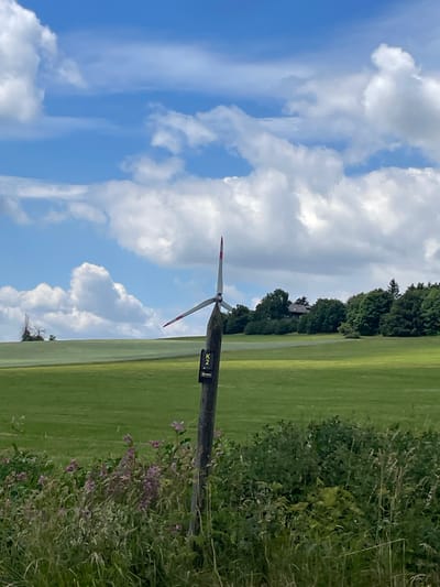 A wind turbine with red and white blades stands in a green field under a blue sky with white clouds. A wooden post with a sign indicating "K 2" and an arrow is in the foreground. In the background, there is a treeline and a building. The scene appears to be located in Schwarzenborn, Germany.