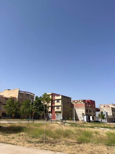 A street scene in El Hajeb, Morocco, captures a row of apartment buildings under a clear blue sky. The buildings, varying in height and architectural style, feature earthy tones with some splashes of red on the roofs and facades. Some of the buildings appear to be under construction or renovation, with exposed brickwork visible on one structure. In front of the buildings, a grassy area with dry patches of yellow grass leads to a paved walkway. Several park benches are scattered throughout this green space, and young trees are planted in the foreground. A few parked cars are visible, including a silver van and a dark-colored vehicle. The overall impression is one of a residential area in a sunny, dry climate.