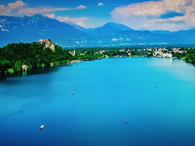 An aerial wide shot captures a vibrant, blue lake bordered by lush green shores and a distant, majestic mountain range. On a rocky outcrop on the left, a castle with red rooftops stands proudly amidst dense foliage. The lake is dotted with several small boats, some with red accents, navigating the calm waters. A cluster of buildings, possibly a town or village, stretches along the shoreline in the middle ground, with a prominent white church spire reaching towards the sky. The background is dominated by the imposing, snow-capped peaks of the mountains, with a clear blue sky overhead punctuated by soft white clouds. This scene does not depict Olișcani, Moldova.