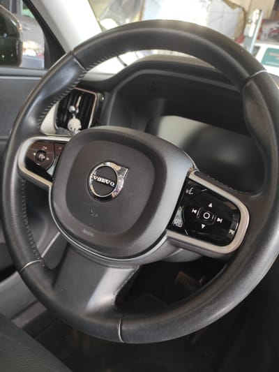 The image shows a close-up of the steering wheel of a Volvo car. The steering wheel is black leather with silver accents. The Volvo logo is prominently displayed in the center of the steering wheel. There are various buttons and controls on the steering wheel, including buttons for volume, track selection, and phone control. A small, decorative charm in the shape of a skull is hanging from the air vent behind the steering wheel. The interior of the car appears to be clean and well-maintained. The image was taken inside the car, likely in a parking lot or garage, as indicated by the background reflections. Without further information, it is not possible to determine the exact location within Saint-Genis-Laval, France.