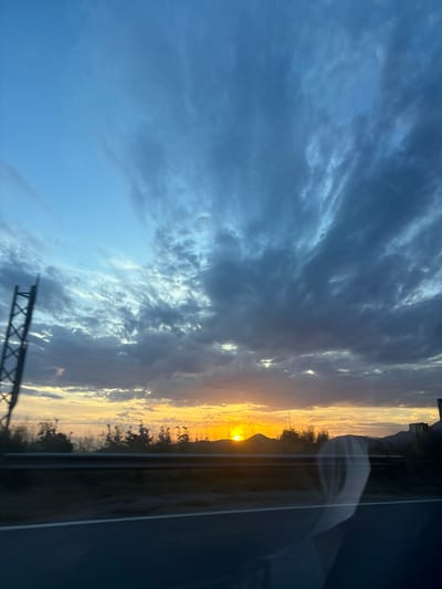The image depicts an outdoor scene at sunset, observed from within a vehicle, likely on a road in the vicinity of Blizikuće, Montenegro. The sky occupies approximately the upper two-thirds of the frame and features a dynamic cloud formation. The upper left portion of the sky is a lighter blue with sparse, wispy clouds, transitioning towards the center and right to dense, dark blue and grey stratiform and cumuliform clouds. These clouds show varying textures and degrees of illumination, with some breaks revealing lighter sky or faint orange/yellow hues from the setting sun.
The sun is positioned low on the horizon, slightly right of the vertical center, emitting an intense orange and yellow glow. It is partially obscured by a dark, silhouetted mountain peak. A range of dark, indistinct mountains and hills forms the distant horizon, with some faint structures visible on the far right.
In the mid-ground, a dark, reflective metal guardrail runs horizontally across the scene. Below the guardrail, a strip of blurred, dark green vegetation is visible. The immediate foreground shows a dark asphalt road surface, blurred due to motion, with a distinct white solid line visible on the right side of the lane.
On the far left, a tall, dark metal lattice structure, likely a utility pole or tower, is partially visible, extending upwards from the lower left corner.
A prominent, partially transparent, whitish reflection of a human figure (head and upper torso) is visible in the lower right quadrant of the image. This reflection, likely from inside the vehicle, overlaps the road and the distant landscape, suggesting the presence of a passenger. The figure appears to have a hood or is wearing clothing that obscures the specific features of the head and shoulders. No specific actions or interactions are discernible beyond the act of observing the sunset from a moving vehicle.