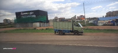 An olive green and blue light truck is traveling on a dirt road in front of a grey and brown commercial building with a green screen. The building has a sign that reads "Ritzuda Coffee". In the background, there are residential buildings and a construction site or junkyard. Utility poles and power lines are visible in the sky, which is overcast with clouds. The text "police kombo" with two heart emojis appears in the bottom left corner.