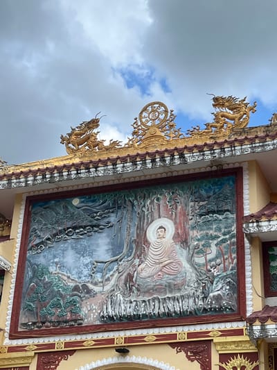 A segment of a temple or pagoda structure in Đăk Mar, Vietnam, is depicted under a partly cloudy sky. The upper portion of the image displays the building's roofline, adorned with intricate, golden, curved decorations. These include two prominent golden dragon statues positioned at the left and right ends of the visible roof section. Centrally located on the roofline is a golden Dharmachakra (Dharma wheel) symbol, flanked by additional golden ornate filigree. The roof features dark-tiled edges and a cream-colored frieze with a repeating pattern.
Below the roofline, a large, rectangular bas-relief sculpture occupies the central area of a cream-colored wall section. This relief features a depiction of Buddha meditating in the lotus position under a large tree, with a bright white halo behind his head. The landscape surrounding Buddha includes stylized mountains, coniferous trees, a body of water, and a full moon visible in the upper left portion of the relief. The relief is framed by a thick, dark red border. The building wall below the relief is a lighter cream color with additional red and golden architectural embellishments.