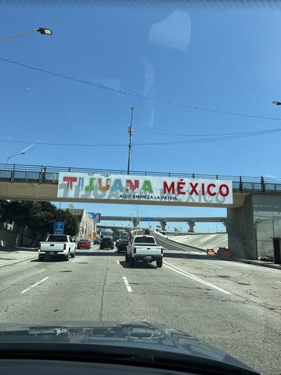 The image shows a street in Tijuana, Mexico, beneath a bridge displaying a sign that reads "TIJUANA MEXICO AQUI EMPIEZA LA PATRIA." Several vehicles are visible on the road, including two white pickup trucks in the foreground. The sky is clear and blue. The photo appears to have been taken from inside a vehicle.