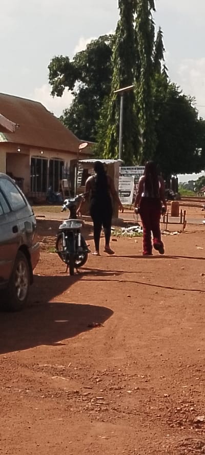The image depicts an outdoor scene in Ajio, Nigeria. Two women are walking away from the camera on a dirt road. The woman on the left wears a black top and dark pants, and the woman on the right wears a white tank top and maroon pants. A dark-colored car is parked on the left side of the frame. In the middle ground, a motorcycle is parked. Behind the motorcycle is a small, open-air structure with signs, and further back, a building with a brown roof and large glass windows is visible. Tall, slender trees with drooping leaves are prominent in the background. The ground is covered in reddish-brown dirt and scattered debris.
