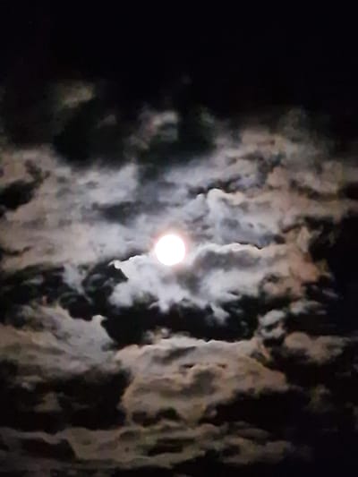 The image depicts a night sky illuminated by a full moon, partially obscured by dark, billowing clouds. The moon is a bright, white orb with a soft glow, positioned centrally within the frame. The clouds are a mix of deep grays and lighter, textured whites, creating a dramatic contrast against the dark expanse of the night. The scene is abstract, with no discernible human figures, man-made structures, or specific landmarks that would indicate a location in Ovoshtnik, Bulgaria, or any other specific geographical area. The focus is solely on the celestial display.