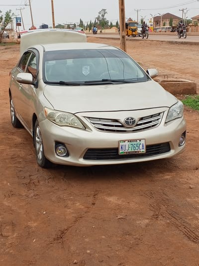 A tan Toyota Corolla is parked on a red dirt shoulder in Badiko, Nigeria. Its trunk is open, revealing a partially visible white object within. The car's license plate reads "KUJ-765CA." In the background, a street scene includes a yellow tricycle (keke), motorcycles, buildings, and power lines. The sky is overcast.
