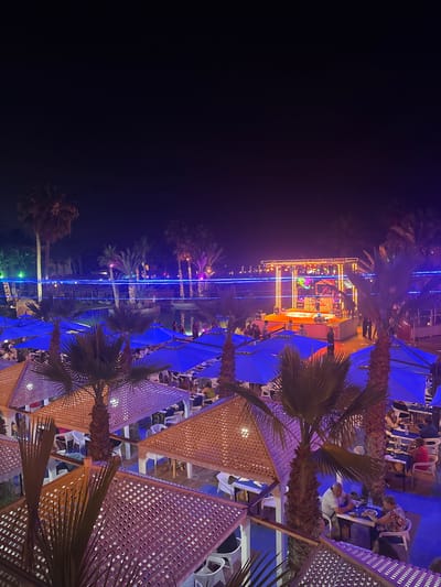 This high-angle aerial view captures an outdoor nighttime venue in Taguermess, Tunisia, featuring a lively atmosphere with a stage, seating areas, and palm trees. A band performs on a brightly lit stage adorned with yellow and orange lights, with musicians visible. In front of the stage, several blue umbrellas shade tables where people are seated, dining and socializing. More seating areas with wooden lattice roofs are visible in the foreground and midground, also occupied by diners. Palm trees are scattered throughout the scene, some with lights strung around them. A swimming pool or body of water is present in the background, with more blue lighting. The overall environment suggests a resort or entertainment complex.