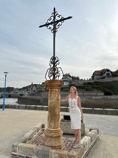 A woman with blonde hair, wearing a white dress with red trim and leopard print shoes, stands outdoors next to a stone pillar topped with an ornate cross. The woman is leaning on the pillar and has her tongue sticking out, looking to her left. The pillar is decorated with carved floral patterns. Behind the pillar and woman is a body of water, a harbor with sailboats, and a hillside with several buildings. The sky is overcast. The scene appears to be located in Saint-Valery-en-Caux, France, on the coast.