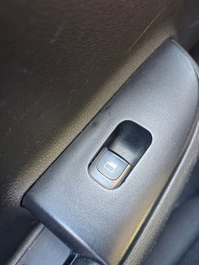 A close-up, eye-level shot focuses on a black car interior, specifically the power window control button on the driver's side door panel. The button is rectangular with a silver-colored outline and a white icon depicting a window with a downward-pointing arrow. It is recessed within a larger, textured black plastic housing. The surrounding door panel is also black with a textured finish, showing some signs of wear, including faint scratches and dust particles. The lighting appears to be from natural daylight, casting subtle shadows and highlighting the textures of the materials. The scene is devoid of any people or overt actions, presenting a static view of a common automotive control. The specific location in Kazachiy Brod, Russia, is not visually discernible from the image itself.