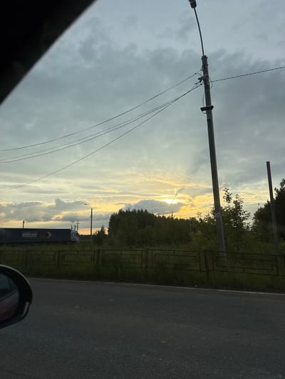 The image captures a roadside scene in Novaya Kazmaska, Russia, likely during the late afternoon or early evening due to the low-angled sunlight. The sky is dramatic, with a mix of dark, ominous clouds and bright, sunlit patches where the sun is breaking through, creating a sense of impending change, perhaps a storm.
Dominating the left side of the frame are utility poles with numerous wires, suggesting an urban or semi-urban environment. A black car's side mirror and part of its frame are visible in the lower right corner, indicating the photograph was taken from inside a vehicle.
The right side of the image features a paved road and a grassy verge bordered by a dark metal fence. Beyond the fence, lush green trees form a dense line, with some taller trees reaching towards the sky. In the background, a dark shipping container with visible white text is situated, along with a small white truck or vehicle. The text on the container includes "CONNECTION" and "LLC," along with other illegible markings. The overall mood is somewhat somber due to the cloudy sky, but the shafts of sunlight add a touch of hope or visual interest.