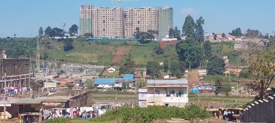 The image presents an elevated, panoramic view of a diverse urban landscape in Limuru Central ward, Kenya, under a clear blue sky.
In the background, on a prominent upper ridge, a complex of several large, multi-story residential buildings is under active construction. These structures feature exposed concrete frames with numerous window openings, and some sections are enveloped in green scaffolding mesh. A yellow tower crane is visible between two of these buildings, indicating ongoing work. To the left of the main construction site, a water tower and communication masts rise above a treed area. Further to the right, additional residential development is scattered across distant hillsides.
The midground features a descending, vegetated slope. A significant electrical substation, equipped with multiple pylons, transformers, and power lines, is situated towards the left-center. Scattered throughout this mid-level are various single and two-story residential buildings, identifiable by their corrugated iron or flat roofs in different colors (blue, brown, white), connected by visible dirt paths.
The foreground displays a dense mix of older and newer structures. On the immediate left, a multi-story building with an exposed concrete frame and laundry hanging to dry suggests a combination of residential and possibly commercial use. Numerous informal or semi-permanent structures with corrugated iron roofs are interspersed. Multiple clotheslines laden with drying garments are visible across the lower residential areas, including near a two-story building with a light blue and white facade. Green shrubbery and a partial gray block wall are present in the lower right. No individuals are distinctly visible, but the extensive laundry and construction activity imply human habitation and ongoing development.
