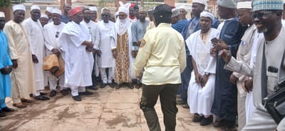 The media file depicts a gathering of men, primarily dressed in traditional Islamic attire, suggesting a cultural or religious event. Several individuals wear white kaftans and caps, with some caps featuring intricate embroidery. One man on the left is wearing a bright blue kaftan. A woman in a light-colored uniform with a black cap stands facing away from the camera, possibly addressing the group or observing the proceedings. The men appear engaged, with some smiling and others looking towards the woman. The setting is outdoors, on a paved area with patches of dirt and scattered debris, possibly in a courtyard or public space. The overall lighting suggests daytime. The presence of a patterned rug being held by one man hints at a ceremonial aspect to the gathering. The men's attire and the relaxed yet attentive atmosphere suggest a community event, possibly a celebration or a meeting of significance. The location context of Sabon Gari North, Nigeria, further reinforces the cultural and social significance of such a gathering. No visible text is present in the image.