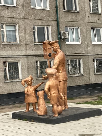 A statue in the city of Usolye-Sibirskoye, Russia, shows a uniformed postman with a mailbag walking with two children and a dog. The statue stands in front of a gray building with multiple windows.
