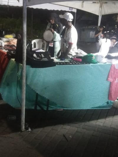 The image depicts an outdoor market stall at night within the Makadara/Mji Wa Kale ward, Kenya. Two individuals are positioned behind a table draped with a light blue tablecloth featuring a scalloped edge. One individual, wearing a white baseball cap and a white short-sleeved shirt with red trim, faces left. The second individual, dressed in dark clothing (possibly including a head covering), holds a light-colored, circular object resembling a plate or bowl, interacting with the first person. A child's face is faintly visible to the left of the table, positioned behind an orange-covered surface.
The table displays various merchandise, including stacks of black t-shirts or bags, one prominently featuring "FREE PALESTINE" text in white. Other visible items on the table include a green cap, a white cap, and a pink cylindrical object, possibly a bottle or container. A white plastic chair is situated behind the table. A white canopy tent structure covers the stall, supported by a light-colored metal pole in the foreground. The ground is paved with dark interlocking blocks. In the background, a dark dumpster-like container is partially visible, and another person in a dark shirt stands near an adjacent white tent. A pink short-sleeved top with white trim hangs from the right edge of the table. The scene is illuminated by artificial light sources, indicating a nighttime setting.