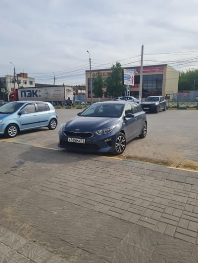 This media file captures a daytime street scene in Novomoskovsk, Russia, under an overcast sky. The image is taken from a slightly elevated perspective, looking down a paved area adjacent to a road.
In the foreground, a dark blue Kia hatchback is prominently parked on a paved surface composed of interlocking grey concrete blocks, with a yellow line marking the edge. The car's Russian license plate, X585 MA 71, is clearly visible. Behind it, a light blue hatchback is also parked. Further along the street, a dark grey SUV is visible.
In the mid-ground, commercial buildings line the street. One building features a large sign with the partially visible Cyrillic text "тная мебель под ключ," indicating a furniture business. Another advertising banner with numbers like "12:4200" and "000 89 19" is also present. Utility poles and a network of power lines crisscross the sky above the street.
The background on the left features a large white truck from the Russian logistics company "ПЭК" (PEK). The truck's side displays the company logo and the text "pecom.ru" and "ДОСТАВЛЯЕМ СБОРНЫЙ ГРУЗ" (Delivering Groupage Cargo). The sky is bright and cloudy, characteristic of an overcast day. The overall scene depicts a typical commercial or urban environment.