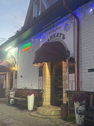 The image shows the exterior of a building in Vereya, Russia, at dusk or early evening. A neon sign reading "ОПТИКА" (Optics) in green and "ОЧКИ ЗА ЧАС" (Glasses in an Hour) in orange is mounted above the entrance. To the right of the optics sign, a larger sign with blue lights and the words "БАНКЕТНЫЙ ЗАЛ" (Banquet Hall) and "АНКЕТЬ" (Anket') is visible. The building has a brick facade and a dark-colored awning over the entrance. A wooden bench with metal scrollwork legs is placed in front of the building, flanked by large white planters filled with purple flowers. A dark-colored trash receptacle is situated near the entrance. The ground in front of the building is paved with cobblestones. The overall scene is illuminated by the building's signs and some artificial lighting, creating a slightly overcast atmosphere.