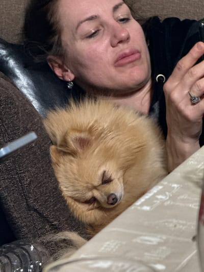 The image depicts a woman lying down with a small, fluffy, golden-brown dog resting on her chest. The woman's face is visible, showing closed eyes. She is wearing a black top with a silver 'C' detail. Her hand is raised, displaying a ring on her finger. The dog is positioned with its head near the woman's neck and its nose visible. A portion of a light-colored, patterned object is also visible in the upper-right corner of the frame. The scene location is not clear enough to provide the city.